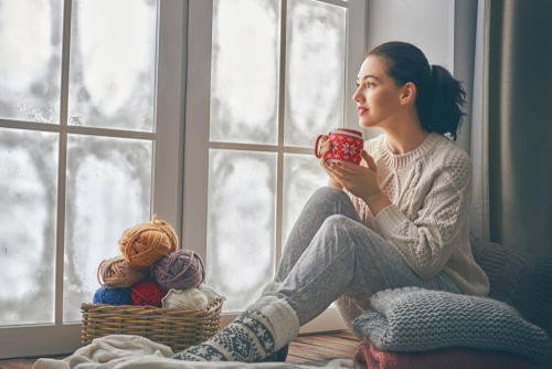 Frau sitzt am Fenster, mit Tasse in der Hand und vor sich ein Korb mit Wolle