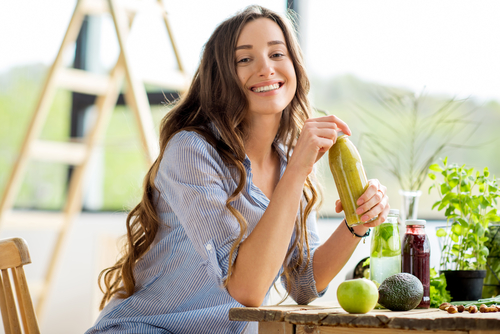 Frau sitzt lächelnd in Küche am Tisch und hält einen grünen Smoothie in den Händen