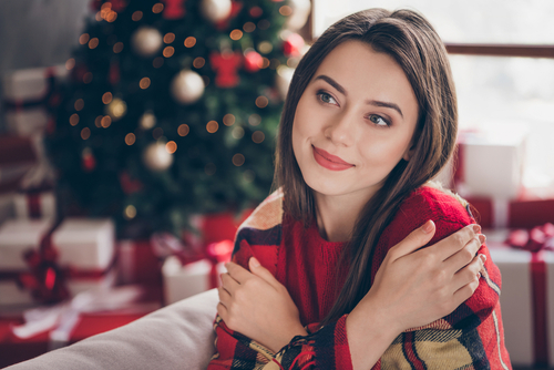 Frau umarmt sich vor Weihnachtsbaum und Geschenken selbst.