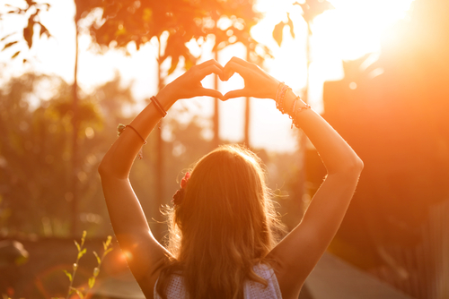 Langhaarige Frau macht mit den Händen ein Herzsymbol während einem Sonnenuntergang in der Natur.